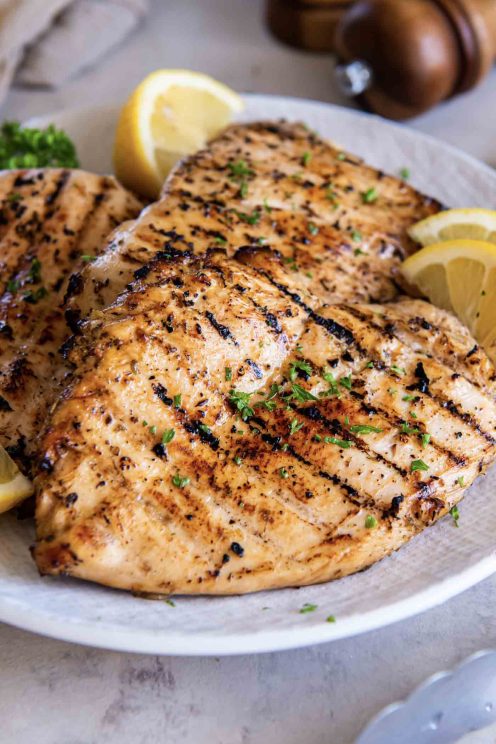 Three grilled chicken breasts on a plate garnished with lemon wedges.