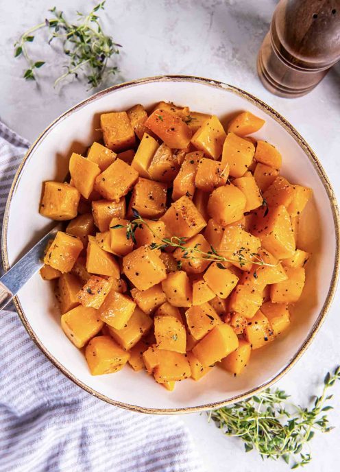 Roasted butternut squash in a serving bowl with a serving spoon, garnished with fresh thyme.