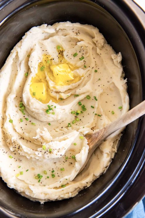 Mashed potatoes in crockpot with butter, pepper, chives and a wooden spoon.