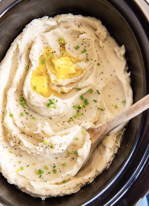 Mashed potatoes in crockpot with butter, pepper, chives and a wooden spoon.