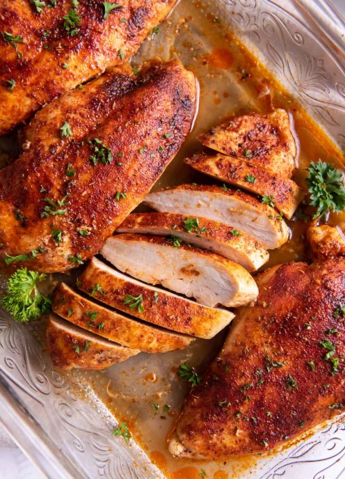 Four baked chicken breasts in baking dish with one breast sliced.