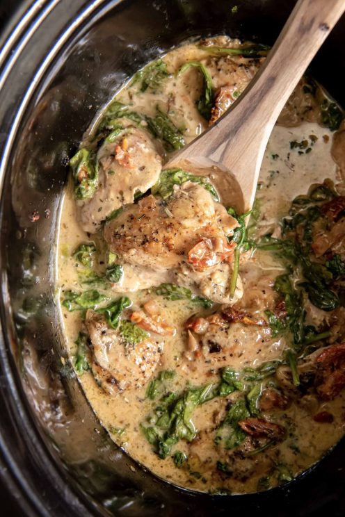 Tuscan chicken thighs in slow cooker with a wooden spoon.