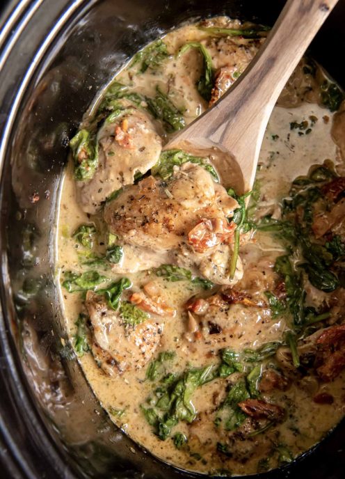 Tuscan chicken thighs in slow cooker with a wooden spoon.
