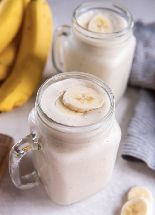 Banana smoothie in a glass topped with two slices of fresh banana with second smoothie glass and bunch of bananas in the background.