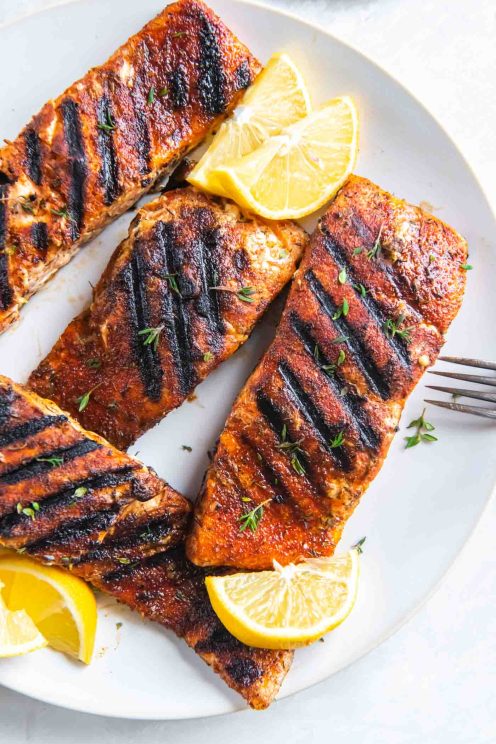 Four grilled salmon fillets on a plate with lemon wedges.