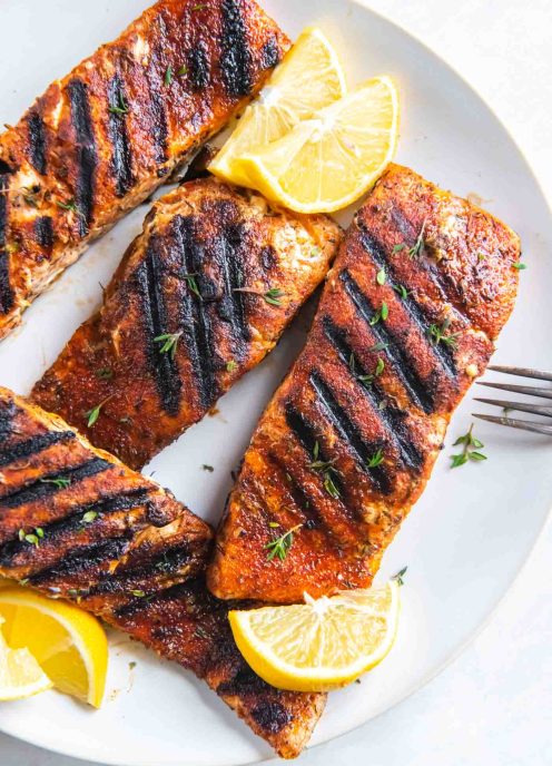 Four grilled salmon fillets on a plate with lemon wedges.