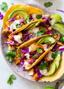 Four fish tacos with red cabbage, avocado, cilantro and fish taco sauce on a plate.