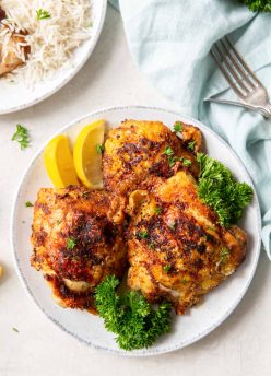 Three crispy bone in air fryer chicken thighs on a plate garnished with lemon wedges and fresh parsley.