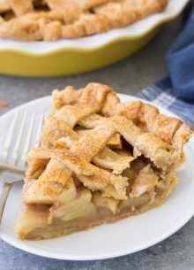 Slice of apple pie on a plate with a fork with whole pie in background.