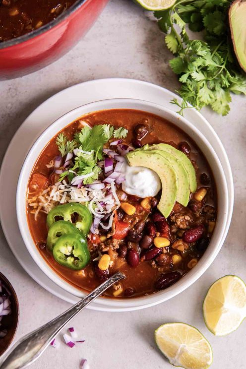 Bowl of taco soup topped with shredded cheese, sour cream, chopped red onion, sliced avocado, sliced jalapeno and fresh cilantro.