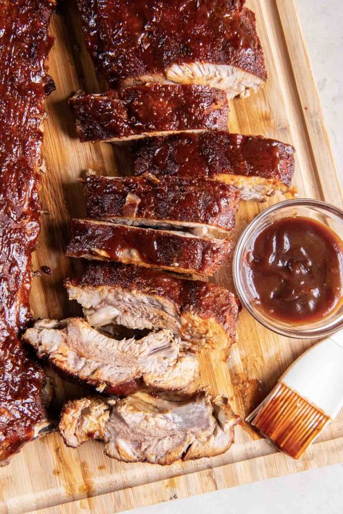 Oven baked baby back ribs on a wood cutting board with a small dish of BBQ sauce.