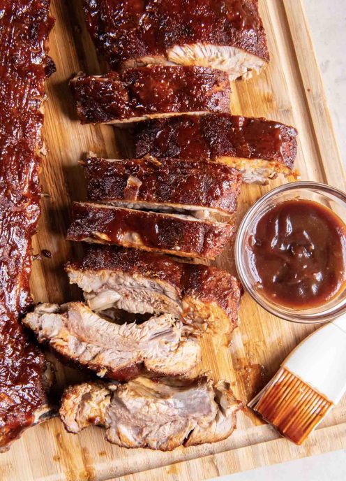 Oven baked baby back ribs on a wood cutting board with a small dish of BBQ sauce.